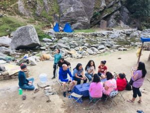 Trekkers sitting around a table near a mountain stream with tents and boulders in the background