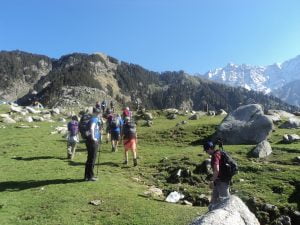 “Trekkers walking through green meadows toward the snow-capped Dhauladhar mountains on the Triund Trek near Dharamshala.”