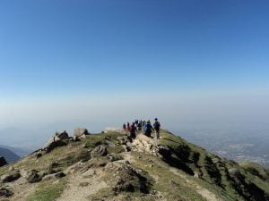 “Trekkers hiking along the ridge of Triund Hill with sweeping views of the valley below and a clear blue sky.”
