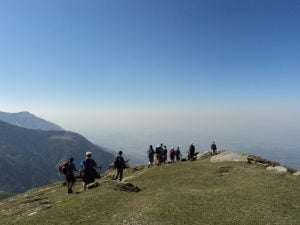 “Trekkers walking across the grassy summit of Triund Top with panoramic views of the Kangra Valley.”