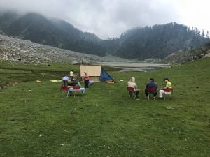 Trekkers relaxing on chairs at Kareri Lake campsite surrounded by green meadows and mountains