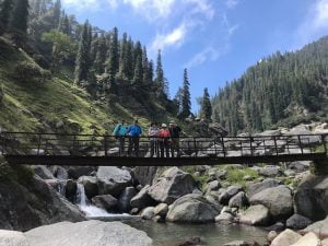 Trekkers crossing a wooden bridge over a rocky stream on the Kareri Lake Trek in Himachal Pradesh with pine forests and mountains in the background