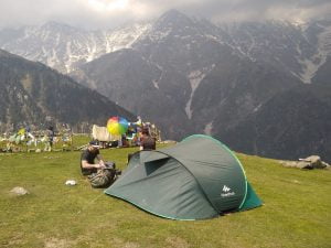 Trekkers resting near tents at Triund campsite with Dhauladhar mountains in the background
