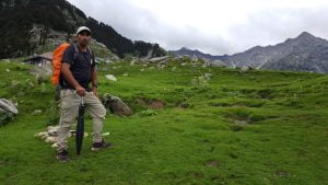 Trek guide standing in a lush alpine meadow with a mountain backdrop in Himachal Pradesh during a summer trek