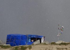 A blue trekking shelter covered with snow beside Tibetan prayer flags at a high-altitude campsite in Himachal Pradesh