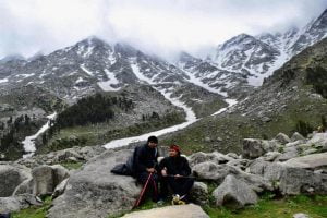 Two trekkers resting on rocks with snow-covered Dhauladhar mountains in the background during Laka Glacier Trek