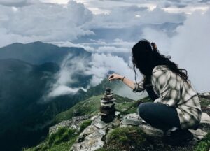 Female trekker stacking stones at a scenic viewpoint on Thatharana Trek with clouds rolling over the mountains