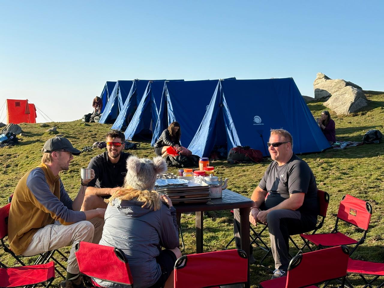 Trekkers sharing a meal beside a blue tent at a mountain campsite