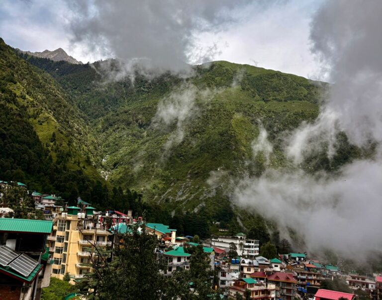 Scenic view of Mcleodganj town with colorful buildings, surrounded by misty green mountains in Dharamsala, Himachal Pradesh.