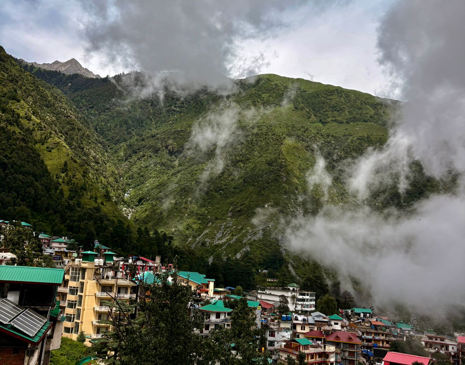 Scenic view of Mcleodganj town with colorful buildings, surrounded by misty green mountains in Dharamsala, Himachal Pradesh.