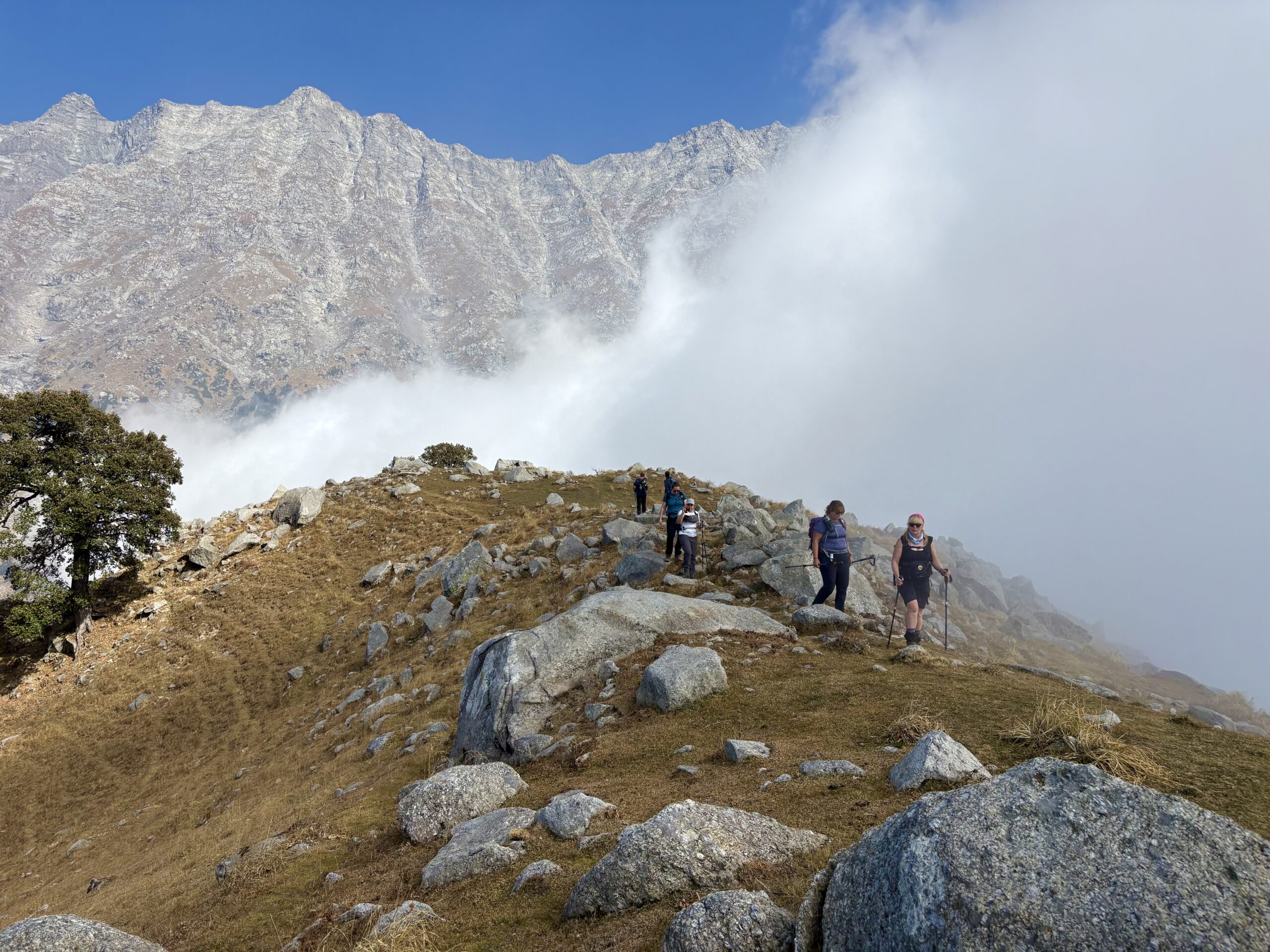 Trekkers walking along a rocky ridge beneath the Dhauladhar mountains and drifting clouds